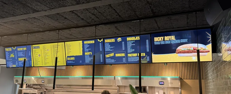 Een lange rij blauw-gele digitale menuborden boven de toog van De Friet Factory, met frieten, snacks, schotels, burgers en een Bicky Royal